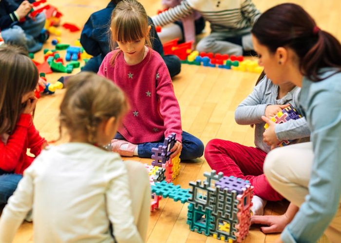 Group of children playing with blocks in kindergarten with their teacher. Showing their imagination, skill and creativity. They are sitting on the floor. Kids are playing and smiling. Teacher is in the middle and helping them to create buildings from blocks.