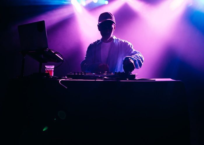 A young African American deejay performs for a crowd at a city night club. Colorful stage lights illuminate the stage behind him.